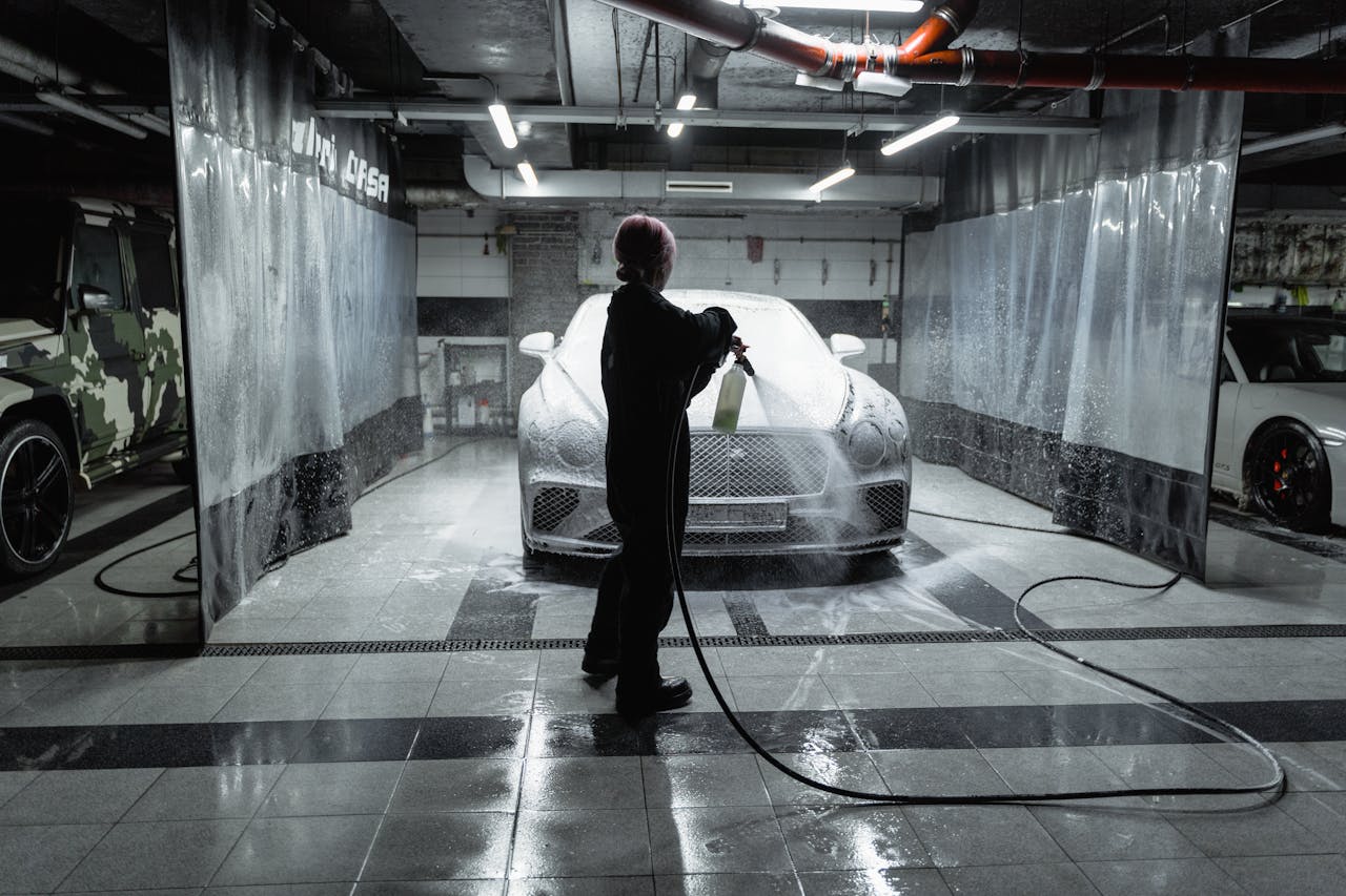 A person using a hose to spray a car with snow foam in an indoor carwash setting.